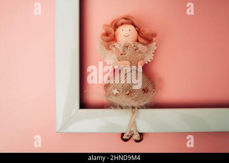 Une figure d'un ange drôle assis sur le bord d'un cadre photo blanc vide sur un fond de pêche, un ange tenant une étoile de Noël Banque D'Images