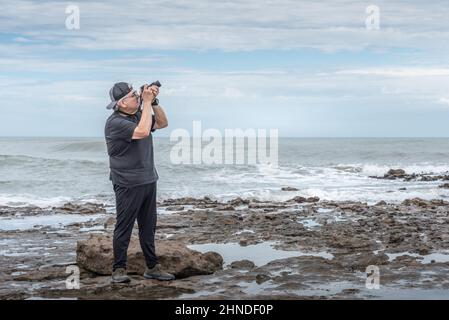 Vue latérale d'un photographe adulte mature avec Cap prenant une photo orientée vers le ciel sur les rochers près de la mer. Banque D'Images