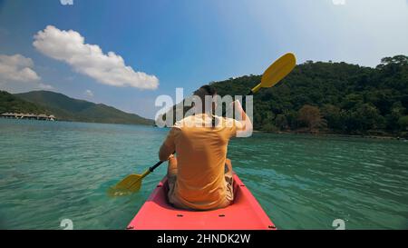 Jeune homme avec des lunettes de soleil et un chapeau rows en plastique rose canoë le long de la mer contre des îles vertes vallonnées avec des jungles sauvages. Voyager dans les pays tropicaux. Un homme fort navigue sur un kayak dans l'océan, vue arrière. Banque D'Images