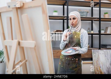 femme arabe pensive avec palette et pinceau pensant près de chevalet à la maison Banque D'Images