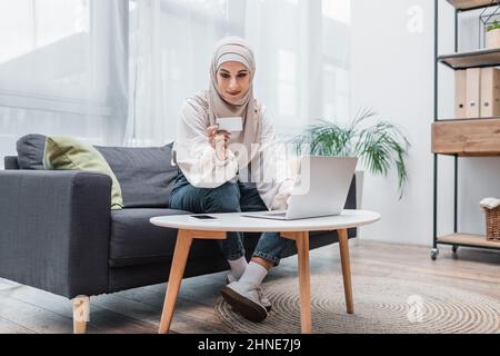 femme arabe souriante regardant la carte de crédit tout en étant assise sur un canapé près d'un ordinateur portable Banque D'Images
