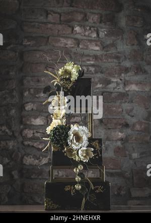 Gâteau de mariage carré à trois niveaux avec fleurs et accents vert foncé sur fond de mur en pierre Banque D'Images