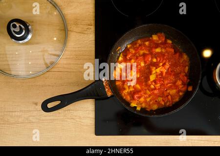 Vue en hauteur de la poêle à frire avec sauce tomate sur une cuisinière électrique moderne, le couvercle en verre se trouve à côté Banque D'Images