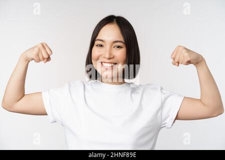 Femme asiatique souriante montrant des biceps flexibles, des muscles forts bras gestuelle, debout dans un t-shirt blanc sur fond blanc Banque D'Images