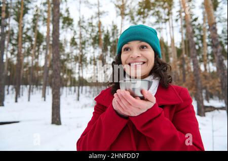 Une charmante femme hispanique vêtue de vêtements chauds tient une tasse thermo avec une boisson chaude près de son visage et sourit avec un magnifique sourire savoureux en appréciant un beau Banque D'Images