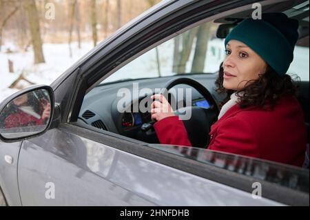 Portrait d'une jolie femme africaine confiante en manteau d'hiver chaud rouge vif et en tricot de laine vert pilote de voyageur, conduite voiture dans une neigeuse W Banque D'Images