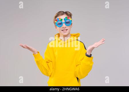 Photo gai excitée jeune gars vêtu d'un sweat-shirt jaune et de lunettes bleues avec l'inscription anniversaire, sourit et fait différents gestes avec ses mains sur les côtés sur un fond gris Banque D'Images