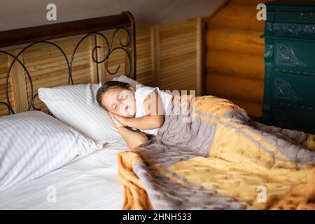 Jeune fille caucasienne prenant la sieste sur un grand lit double avec les deux mains sous la tête recouverte d'une couverture avec la poitrine des tiroirs derrière le lit dans une maison en bois Banque D'Images