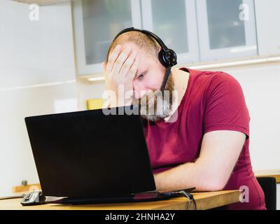 Adulte fatigué tenant la tête avec la main, homme mûr dans le micro-casque travaillant à la maison à l'aide d'un ordinateur portable et fournissant une réunion en ligne, concept de travail à distance Banque D'Images