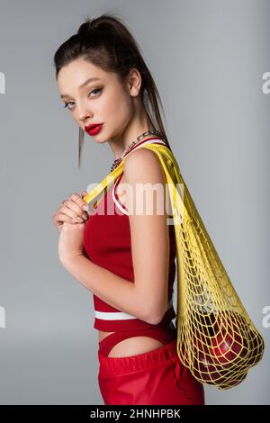 jeune femme élégante tenue sportive rouge tenant un sac à ficelle jaune avec des pommes isolées sur le gris Banque D'Images