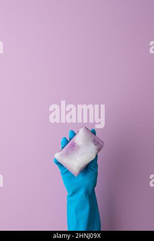 Une main dans un gant de protection en caoutchouc bleu tient une éponge de lavage avec de la mousse sur un fond violet Banque D'Images