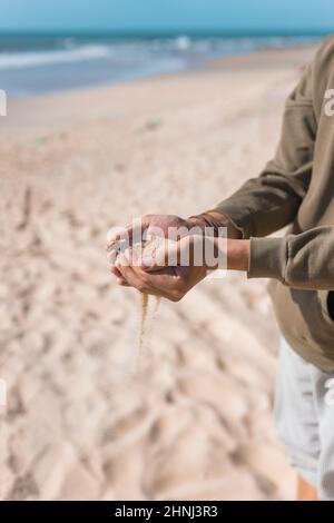 Homme tenant du sable blanc dans ses mains. Palmiers mâles avec le sable sur la plage. Verser le sable. Pull à capuche décontracté. Photo de haute qualité Banque D'Images