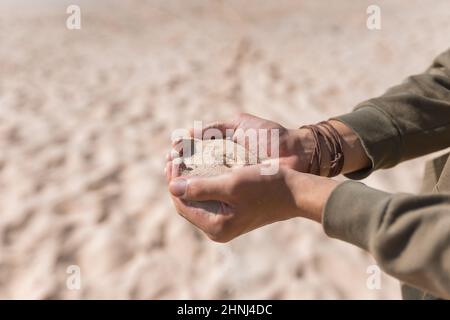 Homme tenant du sable blanc dans ses mains. Palmiers mâles avec le sable sur la plage. Verser le sable. Pull à capuche décontracté. Photo de haute qualité Banque D'Images