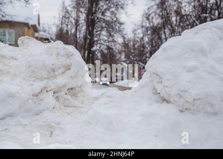 Une grande dérive de neige au bord de la route sur fond de rue de la ville. Sur la route se trouve de la neige sale dans des tas hauts. Paysage urbain d'hiver. Jour d'hiver nuageux, lumière douce. Banque D'Images