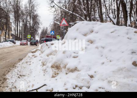 Une grande dérive de neige au bord de la route sur fond de rue de la ville. Sur la route se trouve de la neige sale dans des tas hauts. Paysage urbain d'hiver. Jour d'hiver nuageux, lumière douce. Banque D'Images