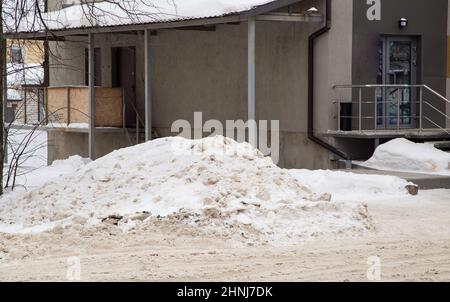 Mars grand glissement de neige par la route sur fond de maisons de ville. Sur la route se trouve de la neige sale dans des tas hauts. Paysage urbain d'hiver. Jour d'hiver nuageux, lumière douce. Banque D'Images