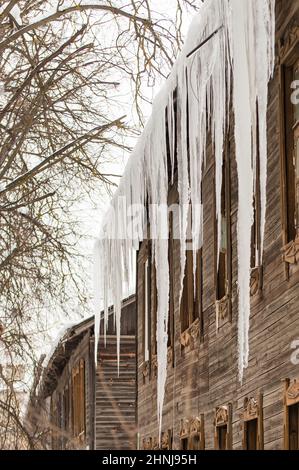 Des glaçons transparents sont suspendus sur le bord du toit. Sur le fond du mur en bois de la vieille maison. Grandes cascades, même de belles rangées. Jour d'hiver nuageux, lumière douce. Banque D'Images