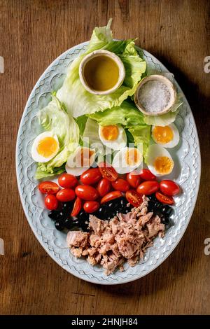 Salade niçoise traditionnelle superposée avec thon en conserve, olives et œufs, servie avec une vinaigrette à l'huile d'olive et sel de mer sur une assiette de céramique sur un bac en bois Banque D'Images