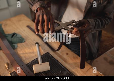 Section médiane de jeunes artisans afro-américains en train de percer un trou sur la ceinture en cuir avec une pince à poinçonner Banque D'Images