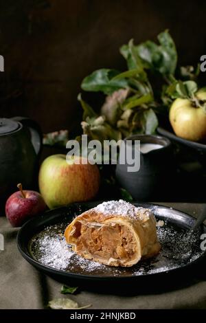 Tranche de tarte traditionnelle maison à la strudel aux pommes dans une assiette en céramique noire servie avec des pommes mûres fraîches, des branches, une fourchette et de la crème sur une nappe en lin sombre Banque D'Images