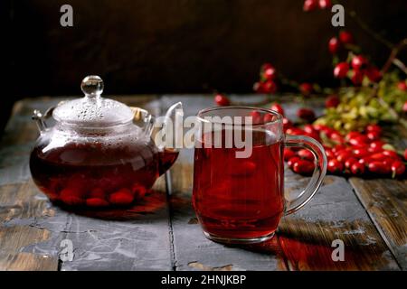 Tasse de fruits de la hanche de la rose tisane et théière en verre debout sur une ancienne table en bois avec baies sauvages d'automne autour.Boisson chaude d'hiver. Banque D'Images