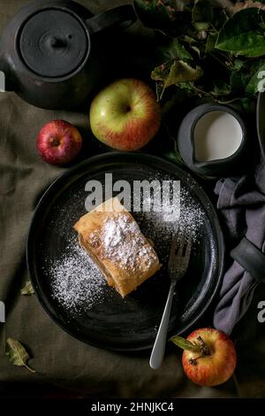 Tranche de tarte traditionnelle maison à la strudel aux pommes dans une assiette en céramique noire servie avec des pommes mûres fraîches, des branches, une fourchette et une théière sur un nappe en lin sombre Banque D'Images