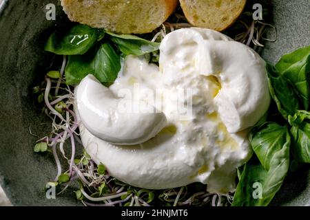 Salade de fromage noué traditionnelle italienne burrata dans un bol en céramique grise sur la table.Pain coupé en tranches, olives, pousses vertes.Dîner méditerranéen sain Banque D'Images