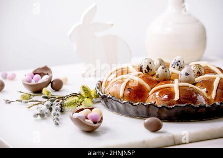 Pains traditionnels de Pâques faits maison à la croix chaude dans un plat en céramique avec des branches de saule en fleur, des bonbons au chocolat et des œufs de caille sur une table en marbre blanc. Pâques Banque D'Images