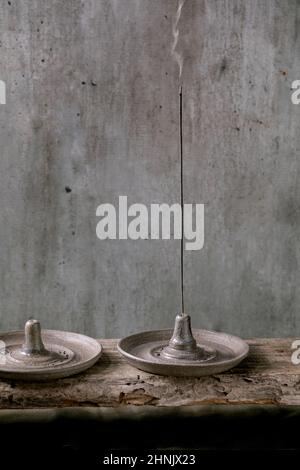 Bâton d'encens fumant dans un porte-encens en céramique gris fait à la main sur une table en bois avec une paroi grise à l'arrière-plan. Banque D'Images