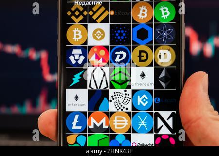 Écran de smartphone avec de petites icônes logos de différentes crypto-monnaies BNB, Binance USD, bitcoin, Cardano, chainlink, COSMOS, Crypto.com pièces, Decentraland, Dogecoin, Electronum, EOS, Ethereum, Huobi Token, IOTA, Kucoin, Litecoin, Monero, Dai, Nano, PROCHE, NEM, Neo, OKB, Polkadot dans une main d'homme, un graphique des statistiques du cours des actions. Février 2022, San Francisco, États-Unis. Banque D'Images