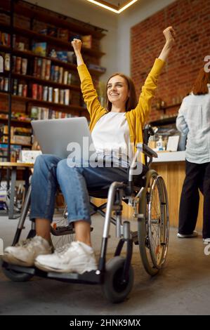 Une étudiante en fauteuil roulant, en fauteuil roulant, en handicap, dans une bibliothèque et à l'intérieur d'une bibliothèque universitaire en arrière-plan. Jeune femme handicapée étudiant à l'université, Banque D'Images