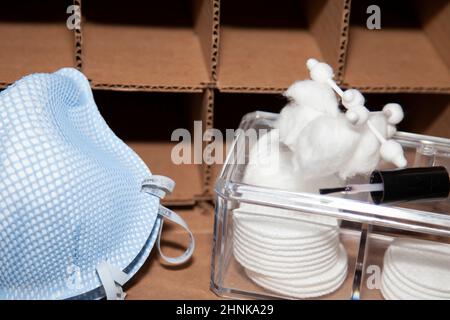 Deux masques N-95 bleus et une boîte en plastique de ronds de maquillage, de boules de coton et de cotons-tiges devant une boîte en carton vide avec des fentes de rangement carrées Banque D'Images