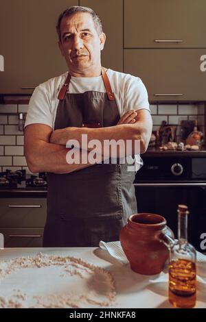 cuisinier d'âge moyen, musclé dans un tablier, bras croisés sur la poitrine, debout dans la cuisine maison. concept de femme de ménage, chef dans la cuisine maison Banque D'Images