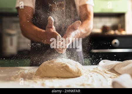 Les mains du chef pulvérisent de la farine sur la pâte. Pétrissage de la pâte. Un chef de cuisine dans le tablier du chef de cuisine vaporise de la farine sur la pâte Banque D'Images