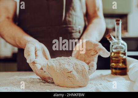 boulanger pour hommes, en tablier brun, pétrir la pâte avec des bras musclés forts, debout dans la cuisine maison. Cuisson de la pâte maison, du pain, des nouilles maison Banque D'Images