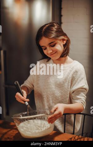 Bonne adolescente cuisinant dans la cuisine, souriante petite fille debout derrière la table chuchotant les œufs et le lait dans le bol tout en préparant la pâte pour le cak fait maison Banque D'Images