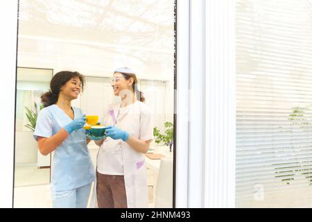 Deux collègues de travail médical multiethniques heureux de sexe féminin qui boivent du café ou du thé et parlent tout en se tenant près de la fenêtre dans une clinique ou un hôpital moderne, t Banque D'Images