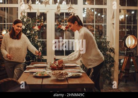 Deux jeunes femmes qui préparent une table de Noël festive à la maison, une mère et une fille heureuses se préparant à la fête de Noël dans une grande ferme rustique et confortable Banque D'Images