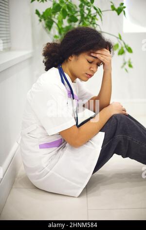 Une jeune infirmière afro-américaine triste et déprimée assise sur le sol avec une expression du visage frustrée, se sentant submergée par la charge de travail en clinique, une femme ethnique Banque D'Images