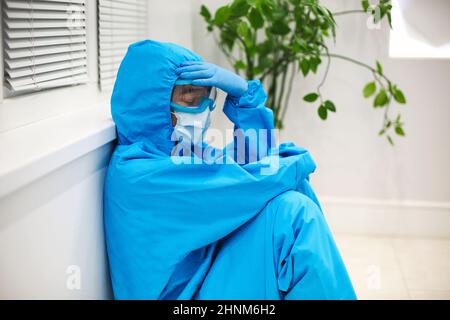 Femme fatiguée médecin dans l'équipement de protection individuelle EPI se sentant déprimé après un déplacement stressant difficile pendant la pandémie Covid-19, assis sur le sol Banque D'Images