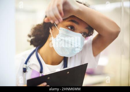 Jeune travailleur médical afro-américain fatigué surpassé portant un masque de protection avec une radiographie dans les mains regardant la fenêtre avec expres frustrés Banque D'Images
