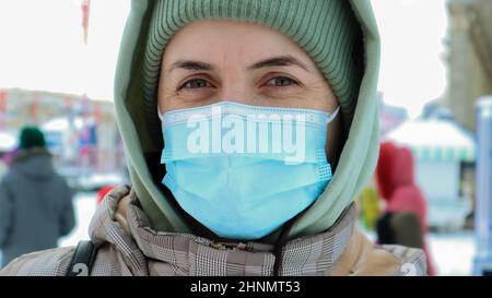 Portrait d'une jeune femme heureuse portant un masque de protection médical à l'extérieur et regardant l'appareil photo. Protection contre les virus. Une femme à l'extérieur par une journée d'hiver se tient dans un chapeau et une cagoule dans le parc. Banque D'Images