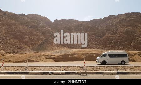 Egypte, Dahab - 17 octobre 2019: Un minibus commercial blanc de passagers avec des touristes sur une route asphaltée dans le désert sur un fond de belles montagnes rocheuses. Banque D'Images