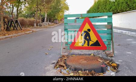 Réparation sur route. Trou d'égout sur la chaussée. Un panneau triangulaire d'avertissement de conducteurs de voiture avec un homme avec une bêche jaune avec rouge. Les hommes au travail. Panneaux de signalisation, restrictions de voyage. Banque D'Images