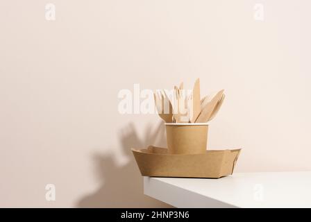 assiettes et tasses en carton brun, fourchettes et couteaux en bois sur une table blanche, fond beige.Vaisselle écologique, sans gaspillage Banque D'Images