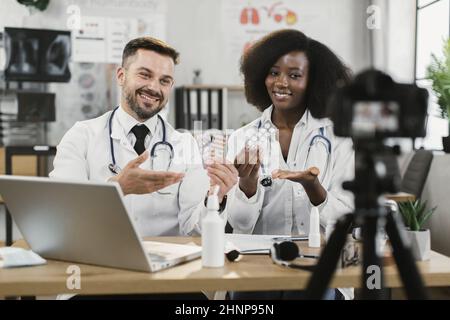 Des médecins afro-américains féminins et caucasiens masculins enregistrent des vidéos sur la prise de médicaments tout en étant assis à leur bureau et en souriant à l'appareil photo. Conseils en ligne de professionnels de santé qualifiés. Banque D'Images