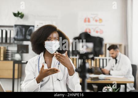 Une femme africaine qualifiée montrant comment porter un masque médical pendant la quarantaine tout en enregistrant un enregistrement vidéo dans la chambre d'hôpital. Concept de la télémédecine, des personnes et de la technologie. Banque D'Images