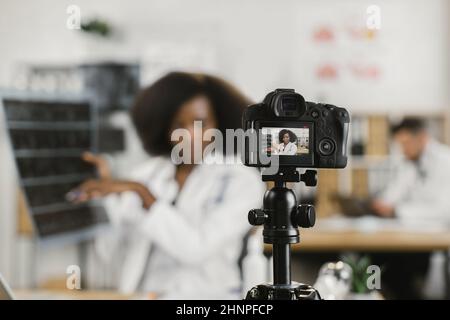 Femme africaine confiante en blouse de laboratoire blanche parlant des résultats de l'acquisition de rayons X pendant l'enregistrement de la diffusion. Mise au point sur l'écran d'une caméra vidéo moderne. Banque D'Images