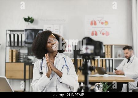 Femme afro-américaine en blouse de laboratoire parlant et faisant des gestes tout en enregistrant un blog vidéo pour sa chaîne médicale. Blog informatif de médecin femme compétent. Banque D'Images