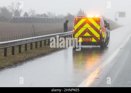 Homme se tenant derrière la barrière de sécurité sur l'autoroute britannique en tant que véhicule de dépannage RAC et mécanicien de travail sur son véhicule en panne en cas de forte pluie - Royaume-Uni Banque D'Images